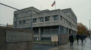 Concrete federal building with a fenced entrance, an American flag, and people walking along the sidewalk