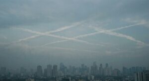 City skyline under a hazy sky with multiple intersecting white vapor trails visible above the buildings