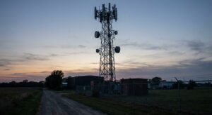 Telecommunication tower with multiple antennas standing near fenced utility buildings at dusk