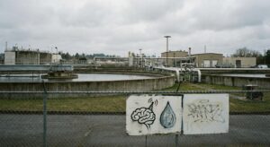 Water treatment facility with circular basins and pipes behind a fence, with a sign showing a brain icon and a water droplet