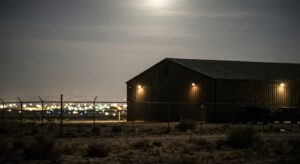 Dimly lit military hangar behind chain-link fencing, photographed at night from outside the perimeter.