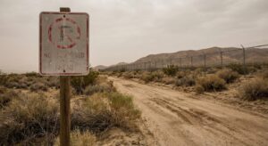 A weathered restricted-area sign beside a dusty desert road with hills in the distance.