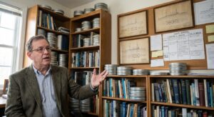 Naval historian in an office with ship diagrams and microfilm canisters while discussing historical records.