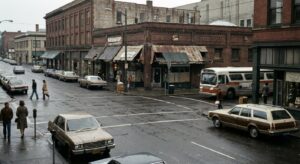 A period-feel Portland street with vintage cars and arcade storefronts, giving historical context to the legend.