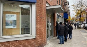 Exterior of a busy bank branch on a city street with customers queued and digital-payment context.