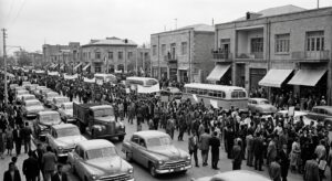 Recreated 1950s Tehran street scene with crowds and vintage cars suggesting a political demonstration.