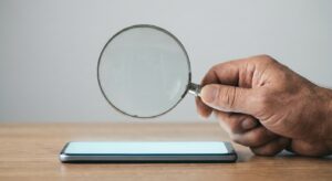 Hand holding a magnifying glass over a smartphone screen, symbolic of consumer scrutiny.