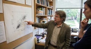 Academic in an office pointing to a large printed scatterplot on a wall during data review.