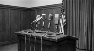 Empty press conference podium with vintage microphones in a plain government hearing room, mid-century aesthetic.