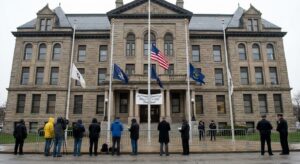 Courthouse façade and flags with neutral onlookers, documentary tone, no text or logos.