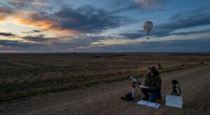 Dusk sky with satellites and a high-altitude balloon visible while a researcher takes notes on the ground.