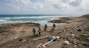 Search teams in protective gloves walking a remote shoreline collecting marine debris into evidence bags.
